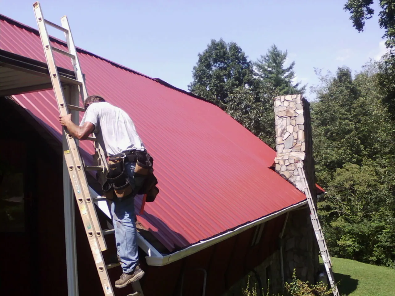 Roofer on a ladder beside a red metal roof and stone chimney — Soffit & Fascia Repair by Roofing Brothers in Stevens Point